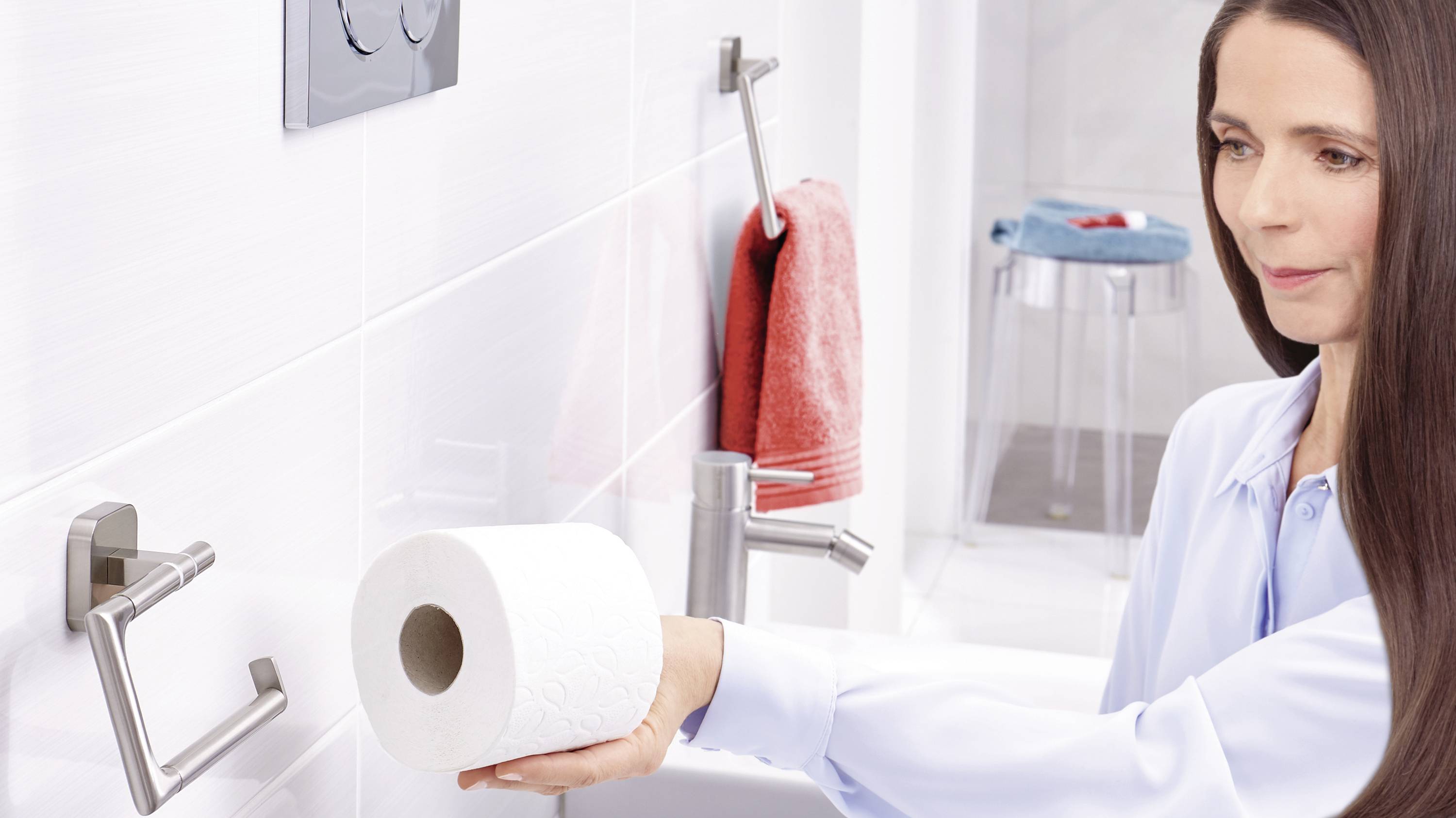A woman is changing a toilet roll in the bathroom. She is wearing a light-coloured shirt and is standing in front of a washbasin with a towel rail.