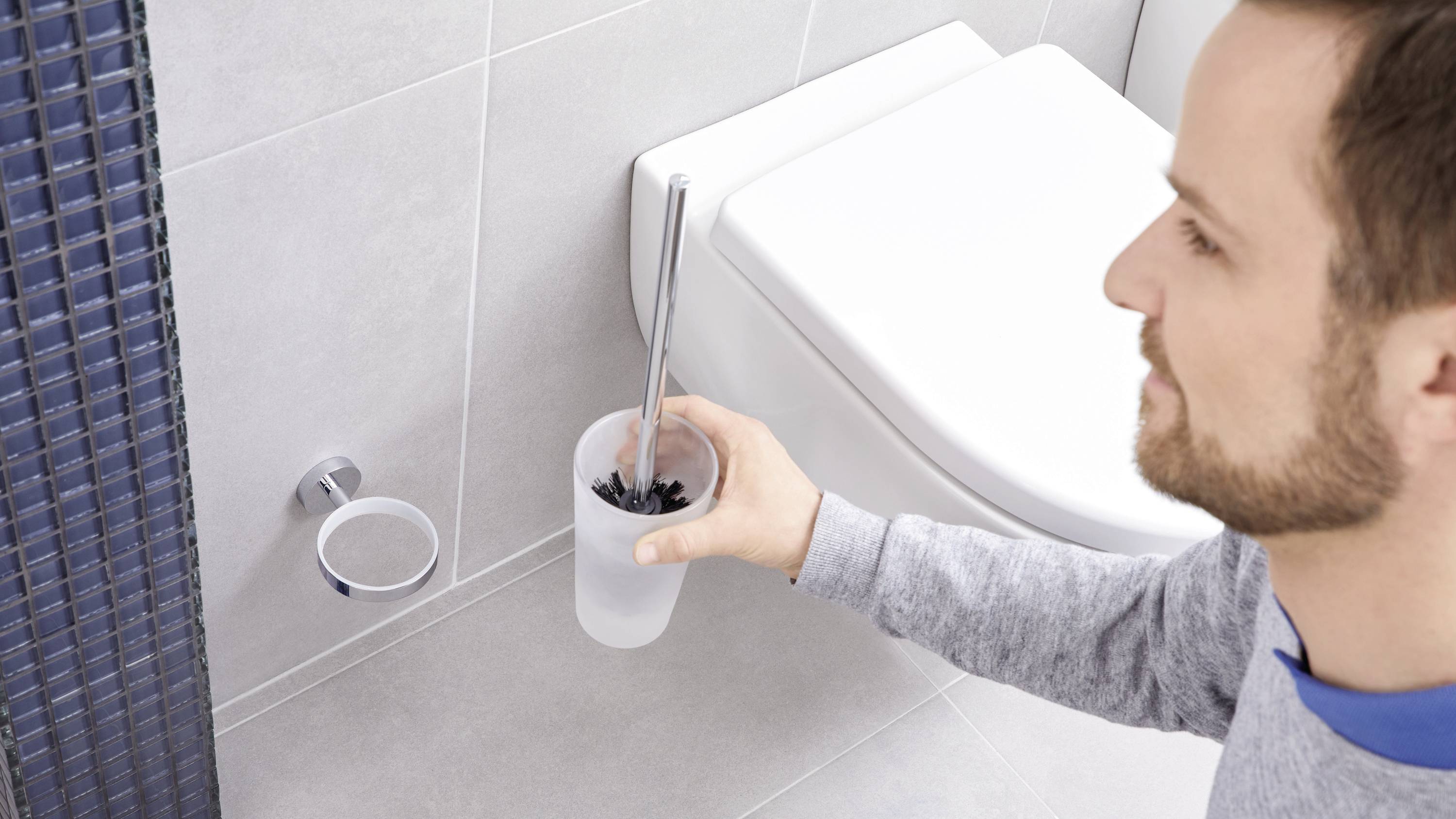 A person is cleaning the toilet with a lavatory brush. A tiled bathroom wall can be seen in the background.