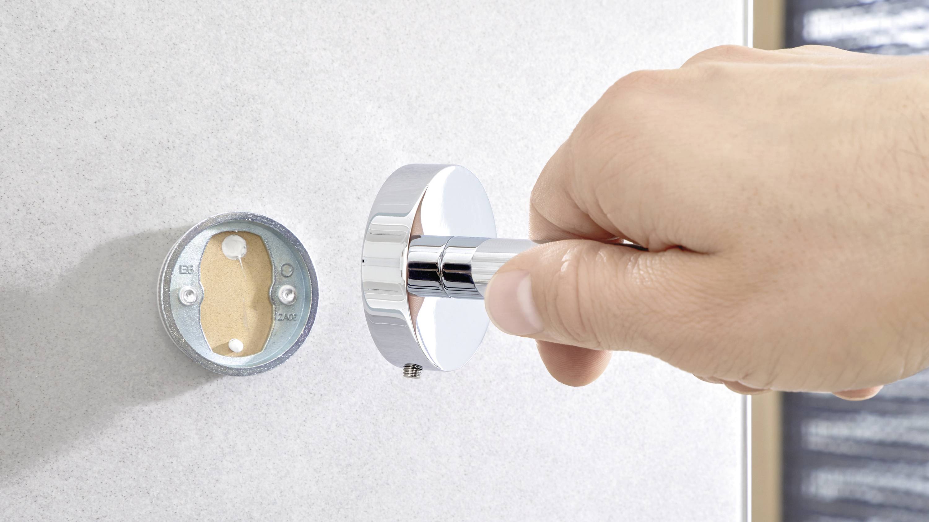 A hand holds a shiny, round door handle and inserts it into a hole in the door, next to an exposed round mounting plate.
