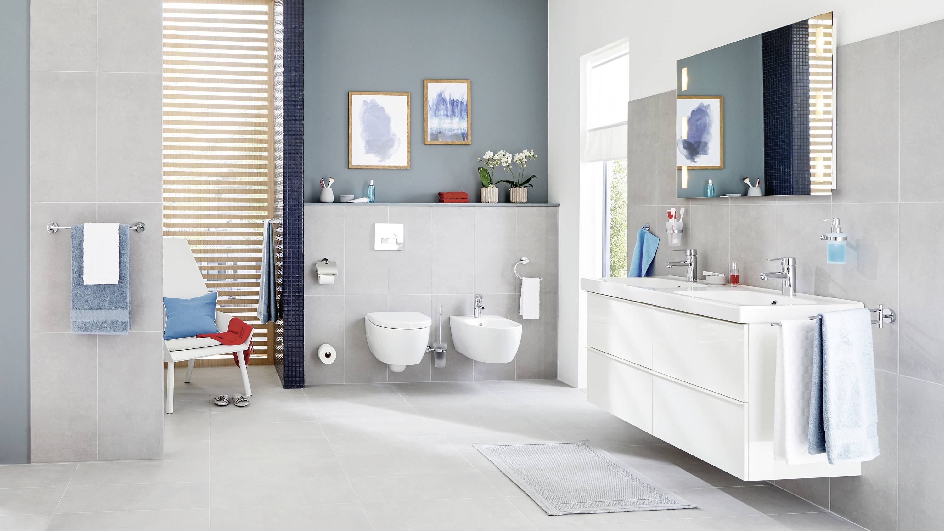 Modern bathroom with grey tiled walls, a wall-mounted toilet, double washbasin with large mirror, decorative plant and artwork on the wall.