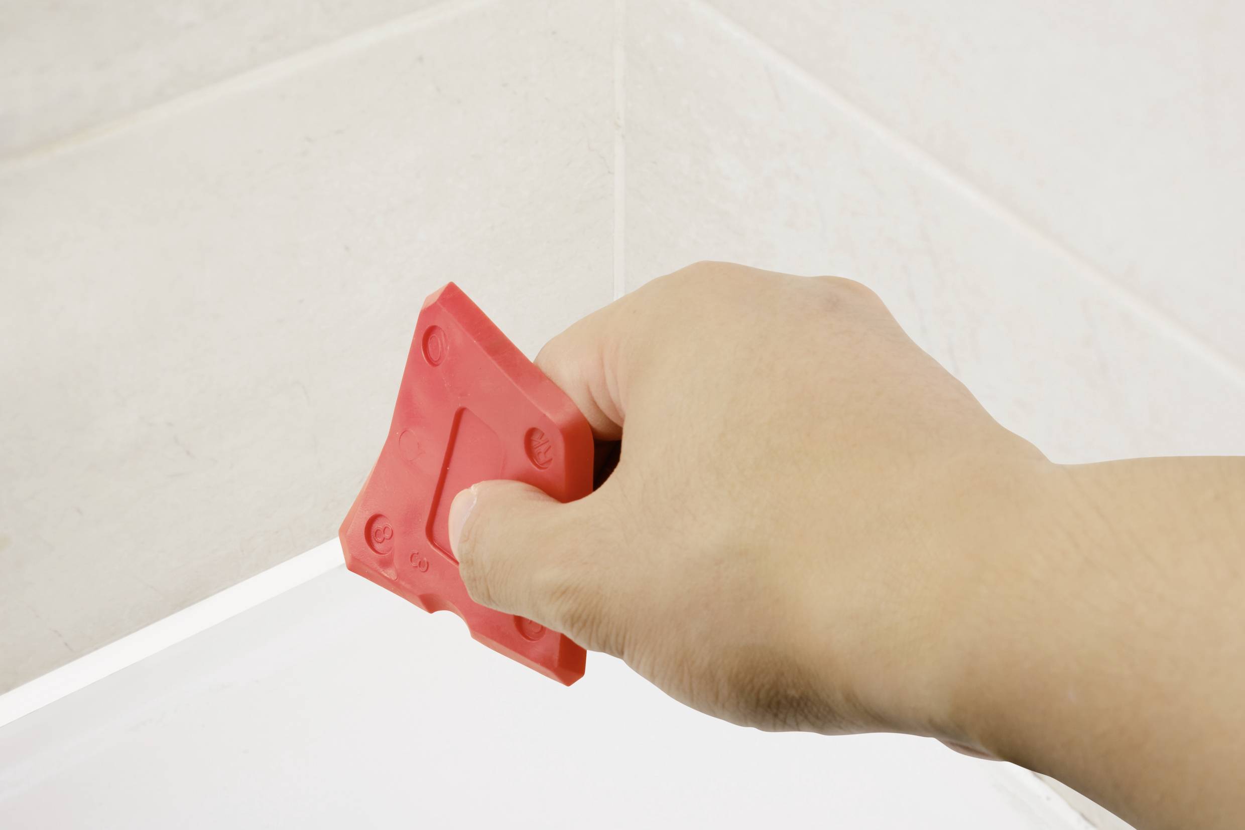 A hand holds a red tool and shapes silicone joints in a bathroom corner.