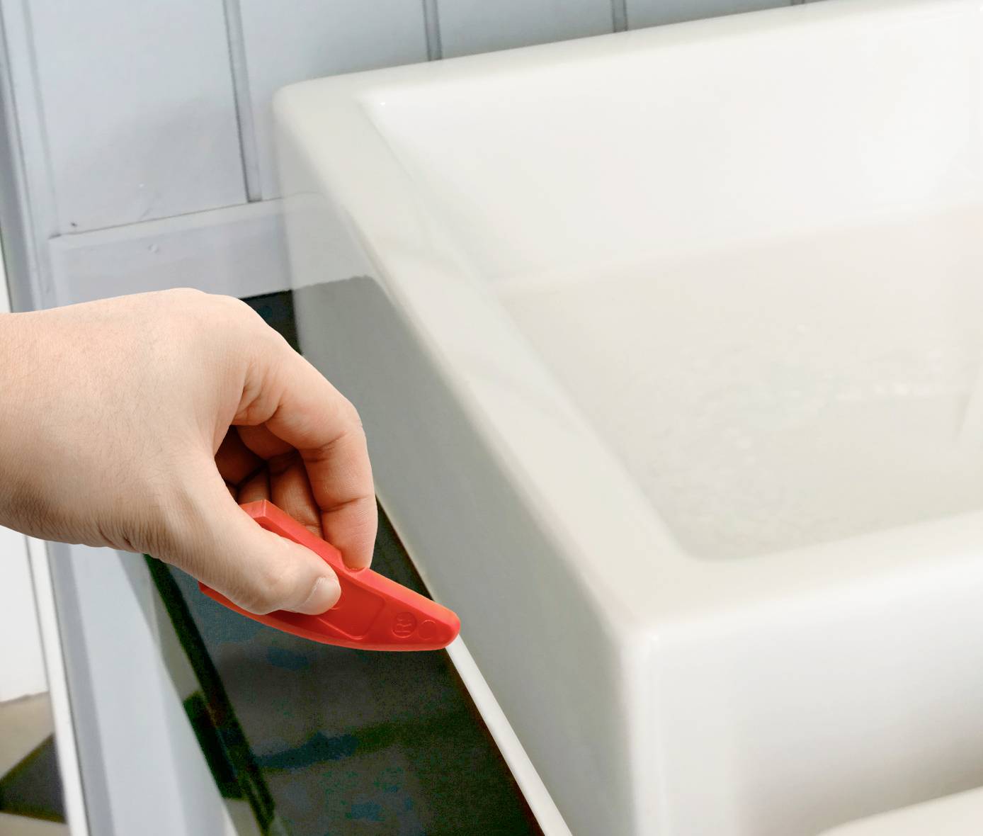 A hand using a red tool to smooth the silicone sealant around a sink.