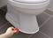 Hand uses a red descaling pad to remove limescale deposits at the base of a toilet on a grey tiled floor.