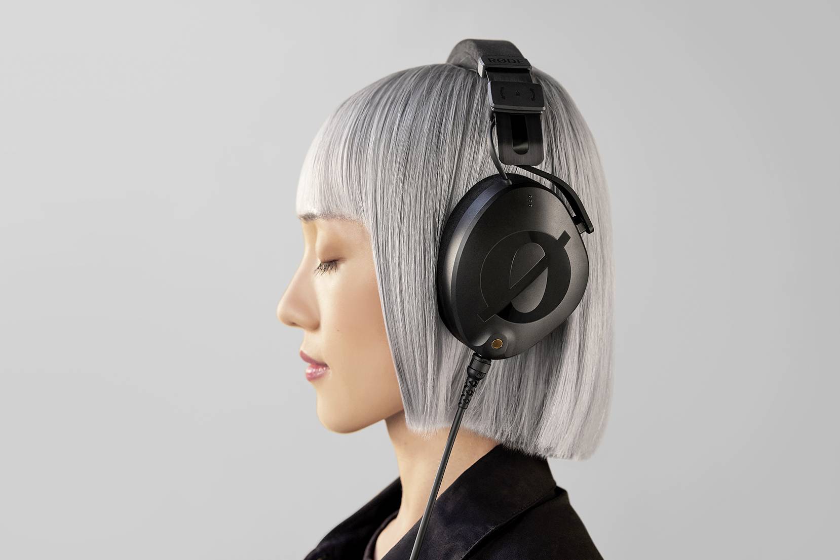 A person with a silver bob hairstyle is wearing large black headphones, viewed from the side profile, against a grey background.