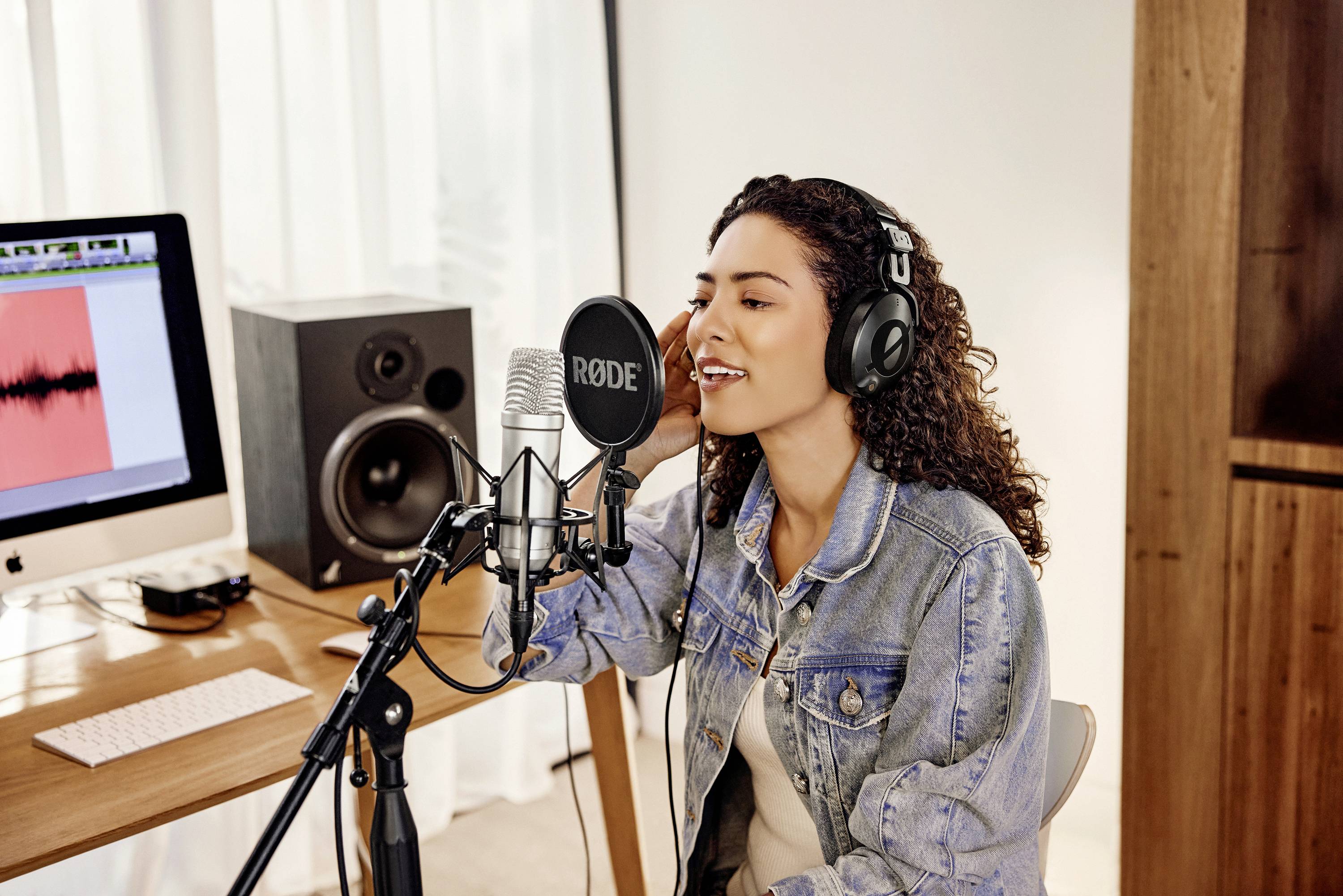 Woman wearing headphones singing into a microphone in a recording studio. In the background, a computer with audio software and speakers.