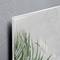 Edge of a glass plate with printed green blades of grass, leaning against a grey concrete wall.