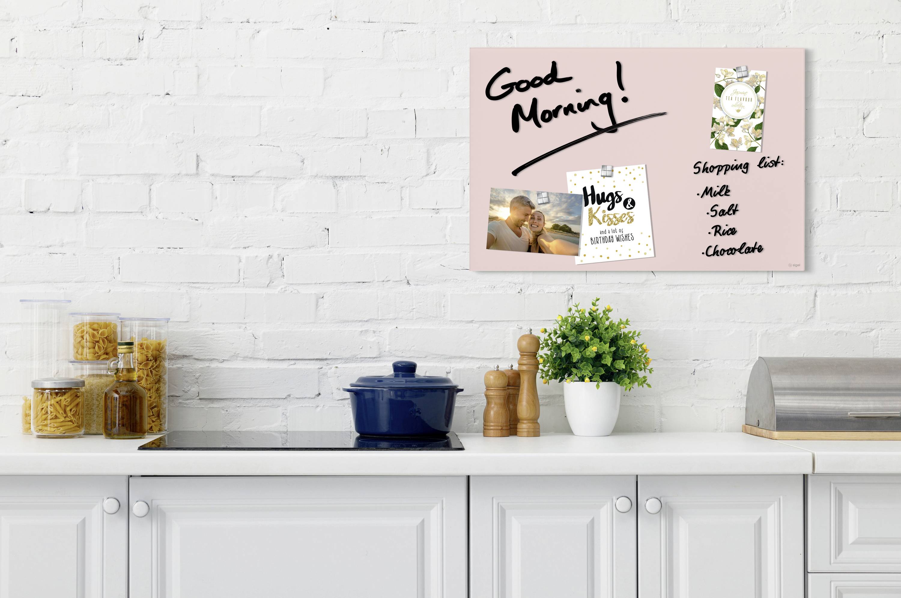 Shelving unit with a pot, wooden utensils and a plant in front of a white kitchen. Pinboard with handwritten 'Good Morning!', photographs and a shopping list.