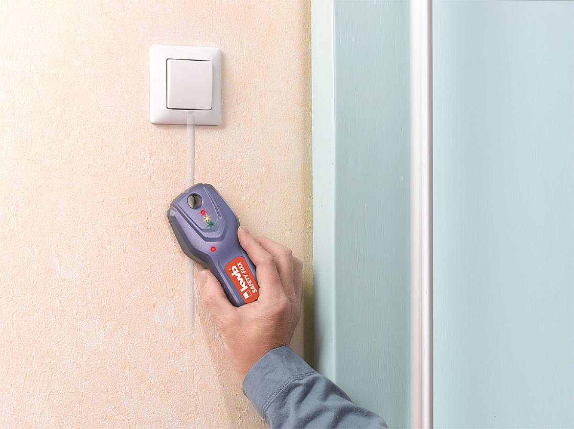 A person is using a digital locator device against a wall to detect pipes and cables behind a light switch.