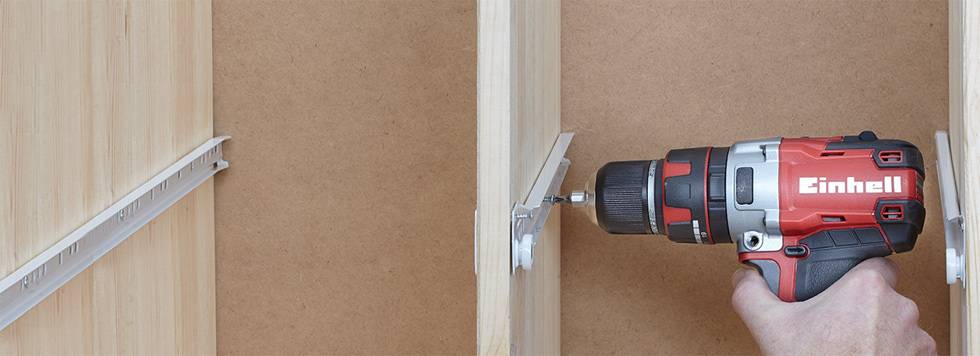 A person is assembling a shelf by turning screws into a brown wooden cabinet with a red cordless drill.