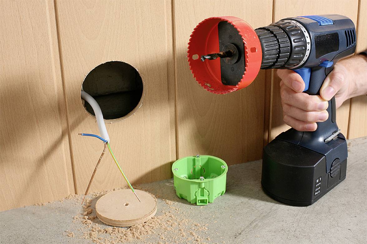 A person is drilling a round hole in a wooden wall using a drill. A green electrical installation box and cables are visible.