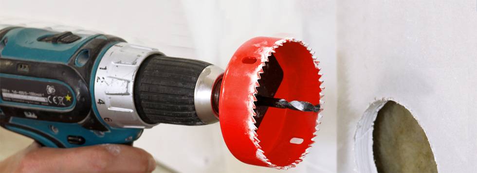 A cordless drill with a red hole saw attachment is drilling a round hole in a white wall.