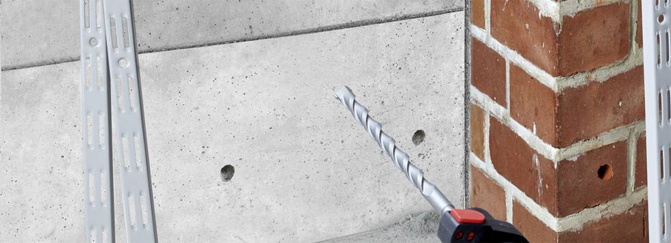 A drill penetrates concrete next to a brick wall. In the background, two metal struts can be seen mounted on the wall.