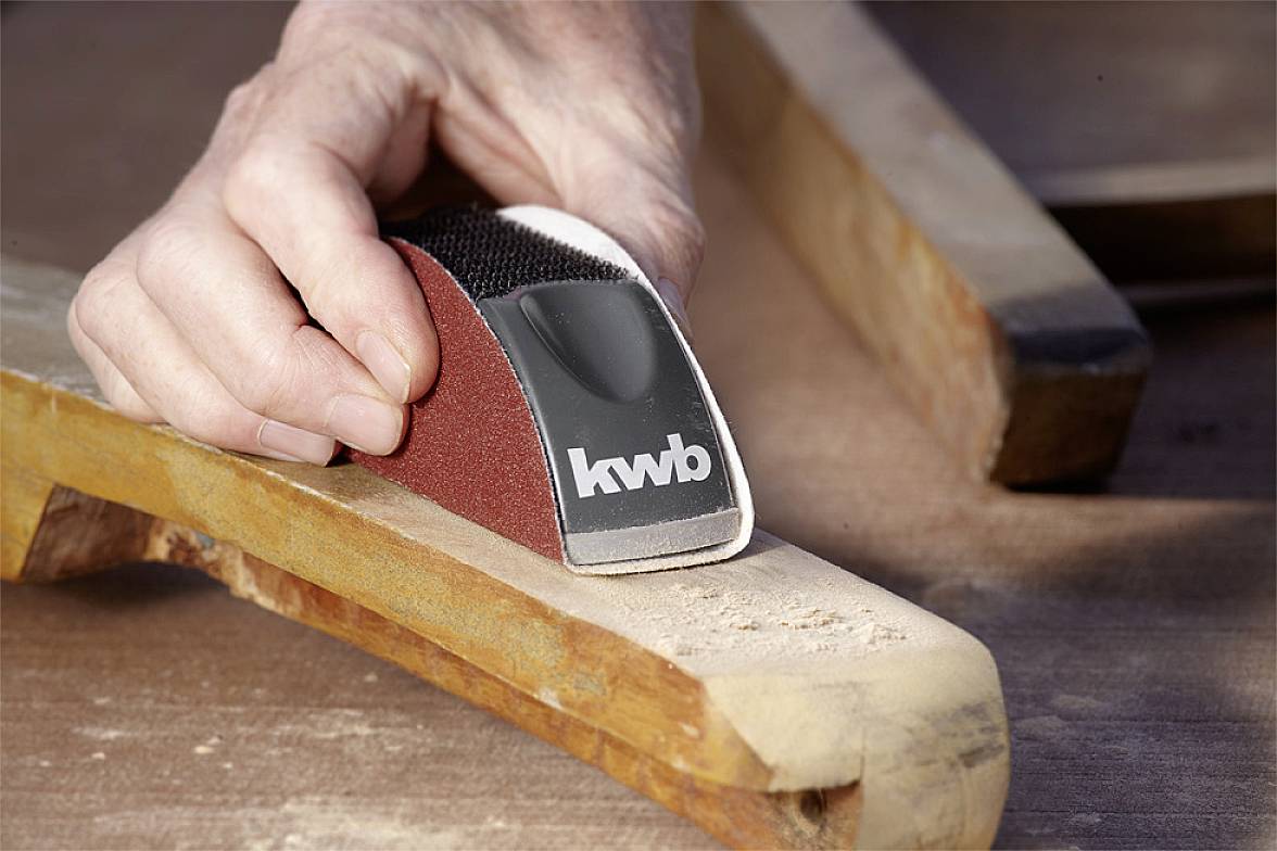 A hand is sanding a piece of wood with a belt sander. The sanding tool bears the 'kwb' logo. Woodworking in action.