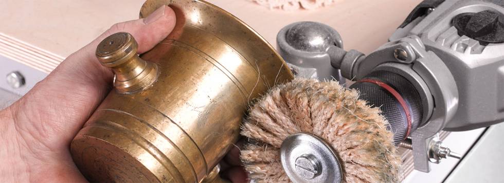 A hand holds a brass vessel that is being polished with a rotating brush. The machine and the vessel are in focus.