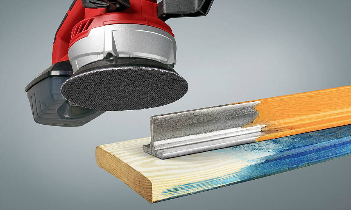 An electric sanding tool removes paint from a wooden board. A piece of metal shows the sanded area.