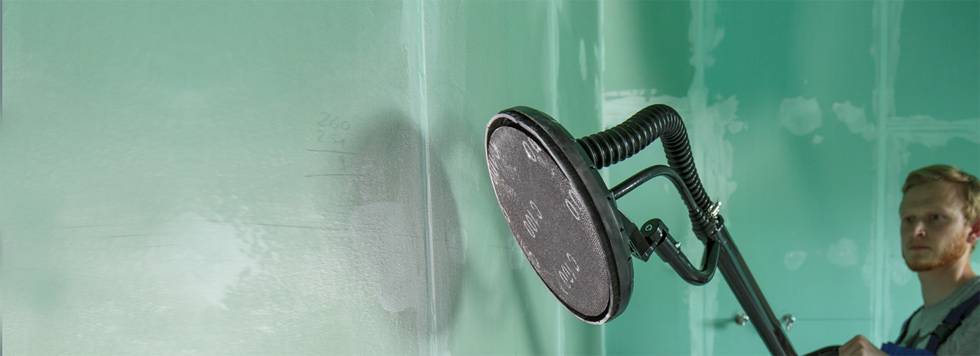 A man is sanding a green wall with an electric sander. He is focused on his work in a brightly lit room.