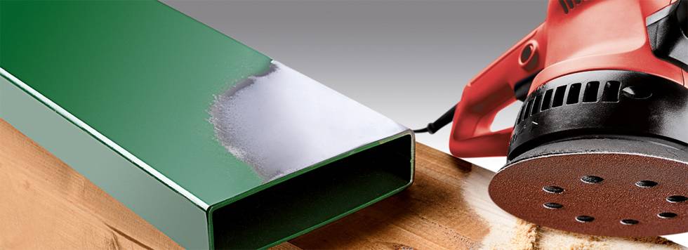A red grinding machine is grinding a green metal bar next to a wooden board.