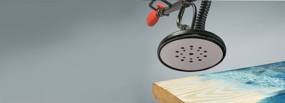 An electric sander with circular sandpaper is working on a wooden tabletop. On the left side of the image, a blue pattern can be seen on the wood.