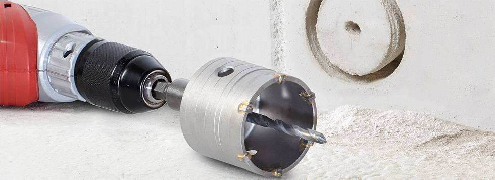 Power drill with hole saw attachment drilling a circular hole in a concrete wall. Dust and debris are visible around the hole.