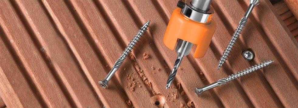 A drill with orange accessories is drilling a hole in a brown, corrugated wooden board, surrounded by four screws.