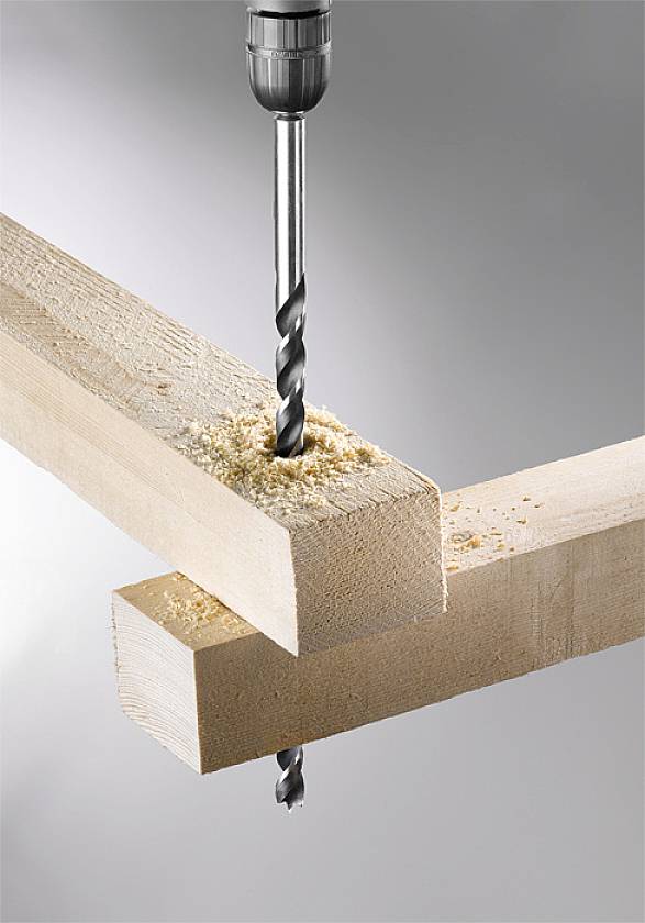 A wooden drill bit bores a hole through two overlapping wooden planks, with wood shavings falling beneath.