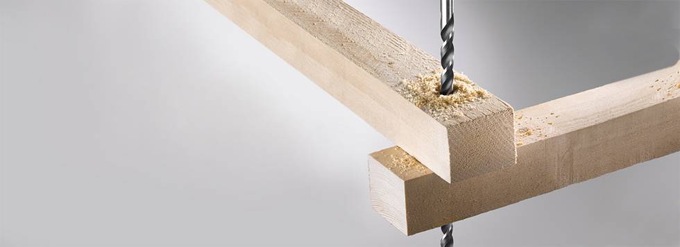 A wood drill precisely bores through two wooden beams lying at right angles to each other, creating sawdust.