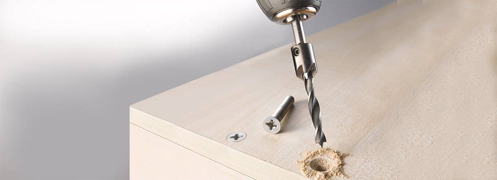 A drill is boring a hole into a wooden board. A screw and an already secured screw can be seen beside it.