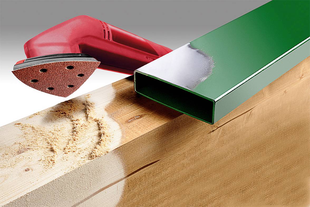 A red sanding block is working on a wooden surface with wood shavings. A green, rectangular metal pipe lies beside it.