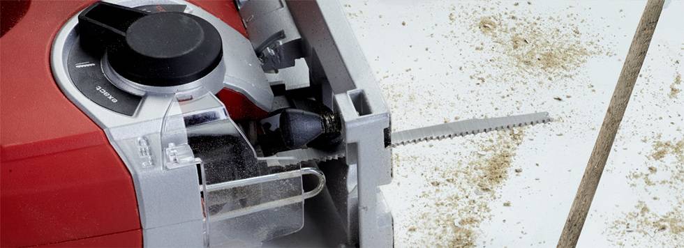 Close-up of an electric jigsaw, blade pointing to the right. Sawdust is scattered around the tool, indicating recent use.