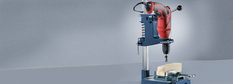 A red drill is mounted on a blue pedestal drill. A piece of wood is being drilled underneath.