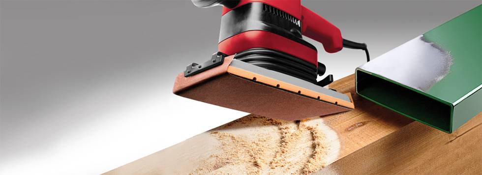 An electric sander smooths a wooden surface, surrounded by sawdust, next to a green metal area.