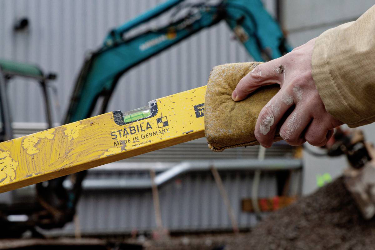 A person is holding a yellow spirit level in front of a construction site with a digger in the background. The spirit level bears the 'Stabila' marking.