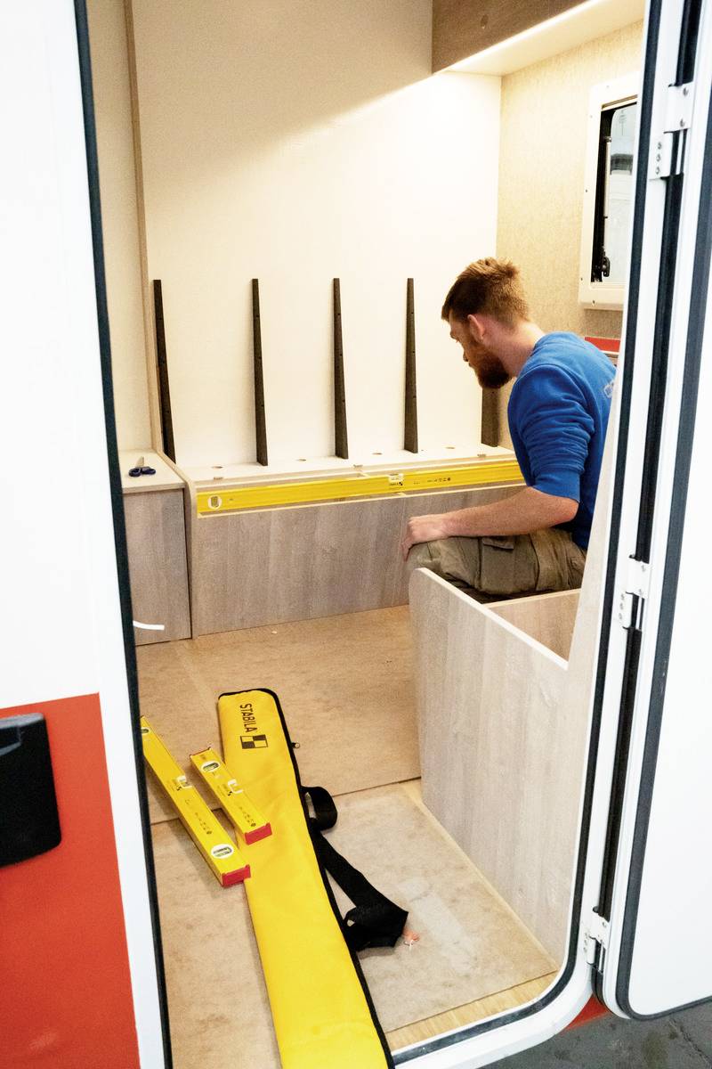 A person is working inside a motorhome interior, surrounded by yellow tools. The space shows unfinished furniture or panelling.