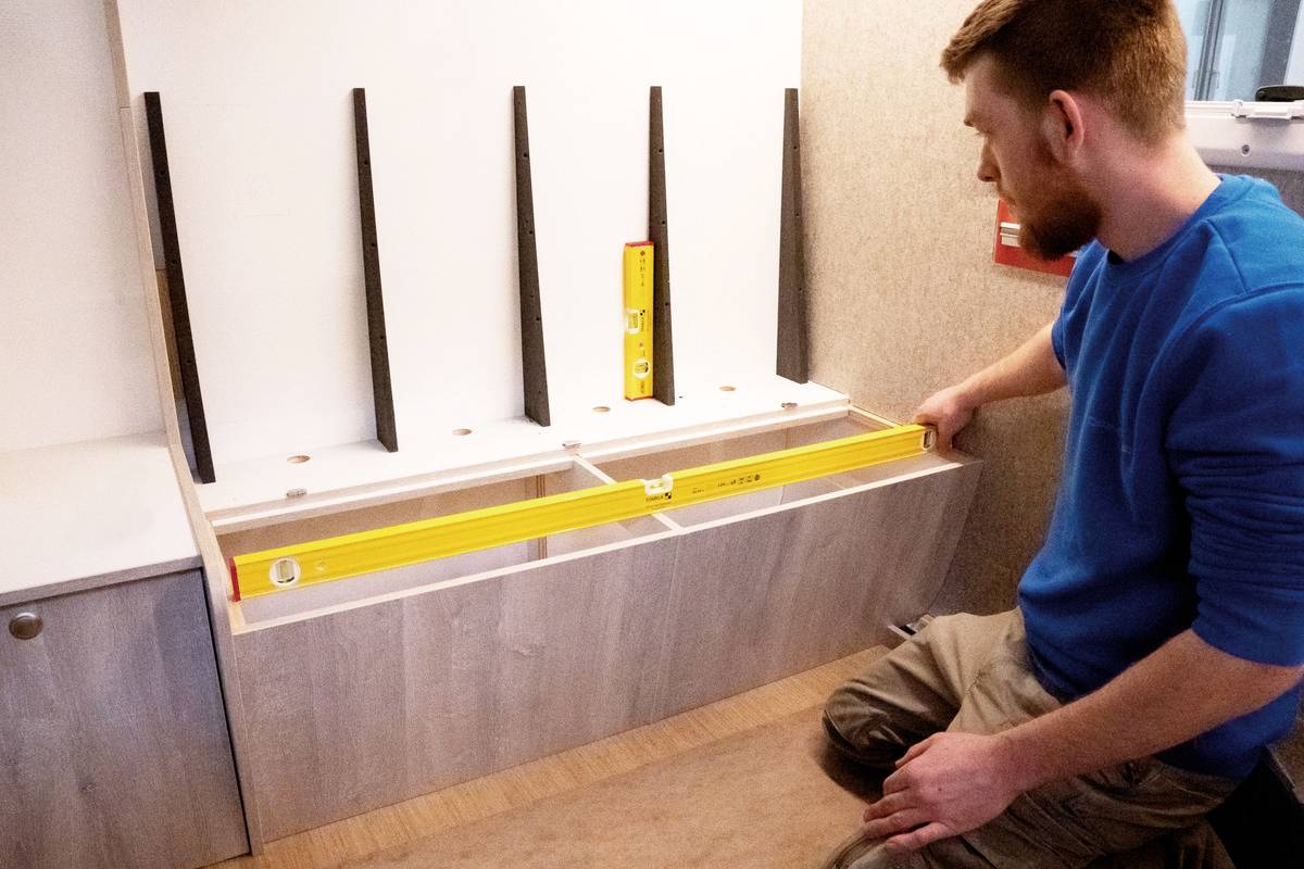 A man in a blue jumper is using a spirit level to check the horizontal alignment of a piece of furniture with white shelves.