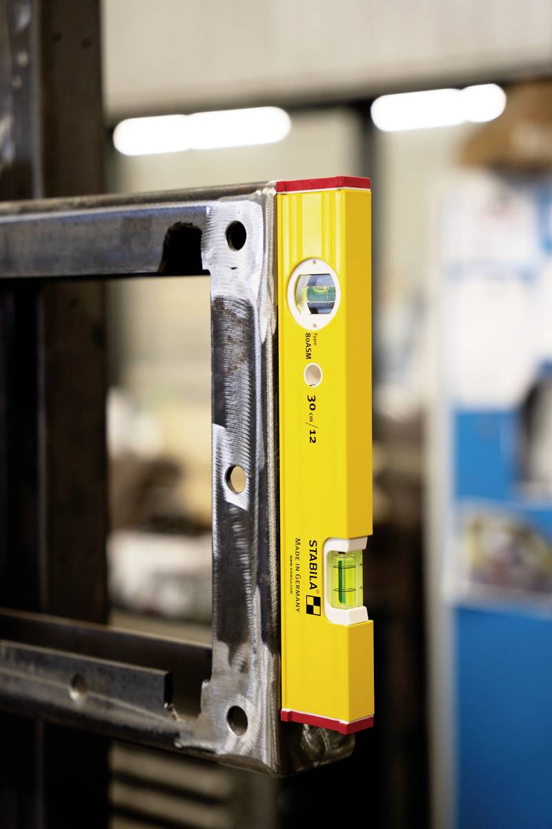 A yellow spirit level indicates a horizontal alignment on a metal frame. Blurred work materials can be seen in the background.