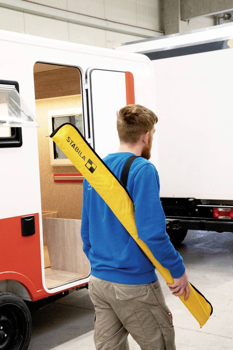 A man in a blue jacket is standing in a workshop next to an open caravan, carrying a yellow bag with the 'STABILA' label.