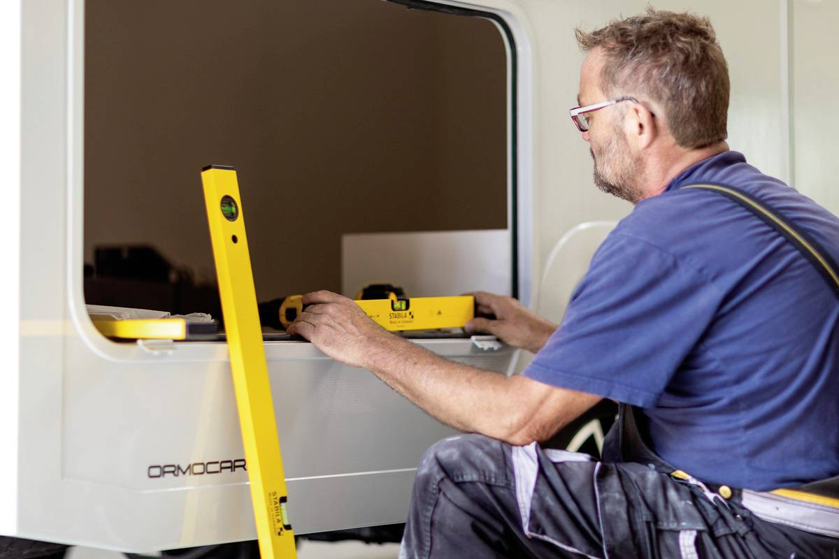 A man is working with a spirit level on a vehicle window frame. He is wearing workwear, with a workshop in the background.