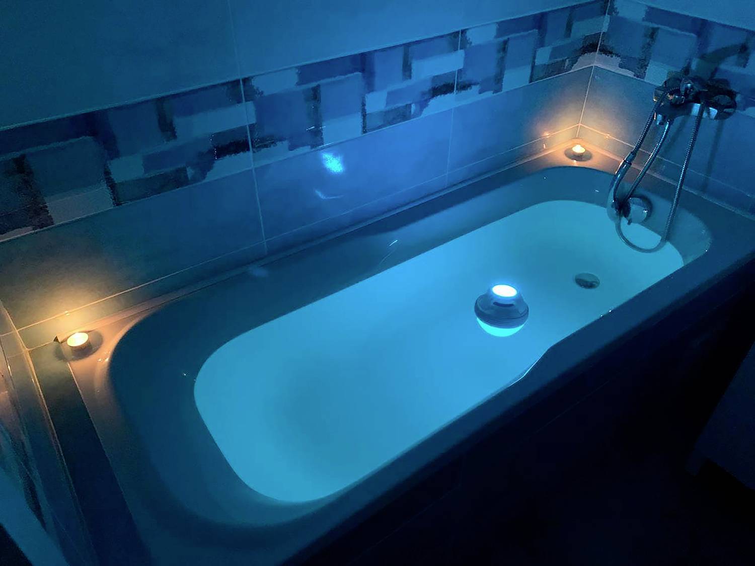 A filled bathtub illuminated by soft, blue light with candles around the edges creates a relaxing atmosphere.