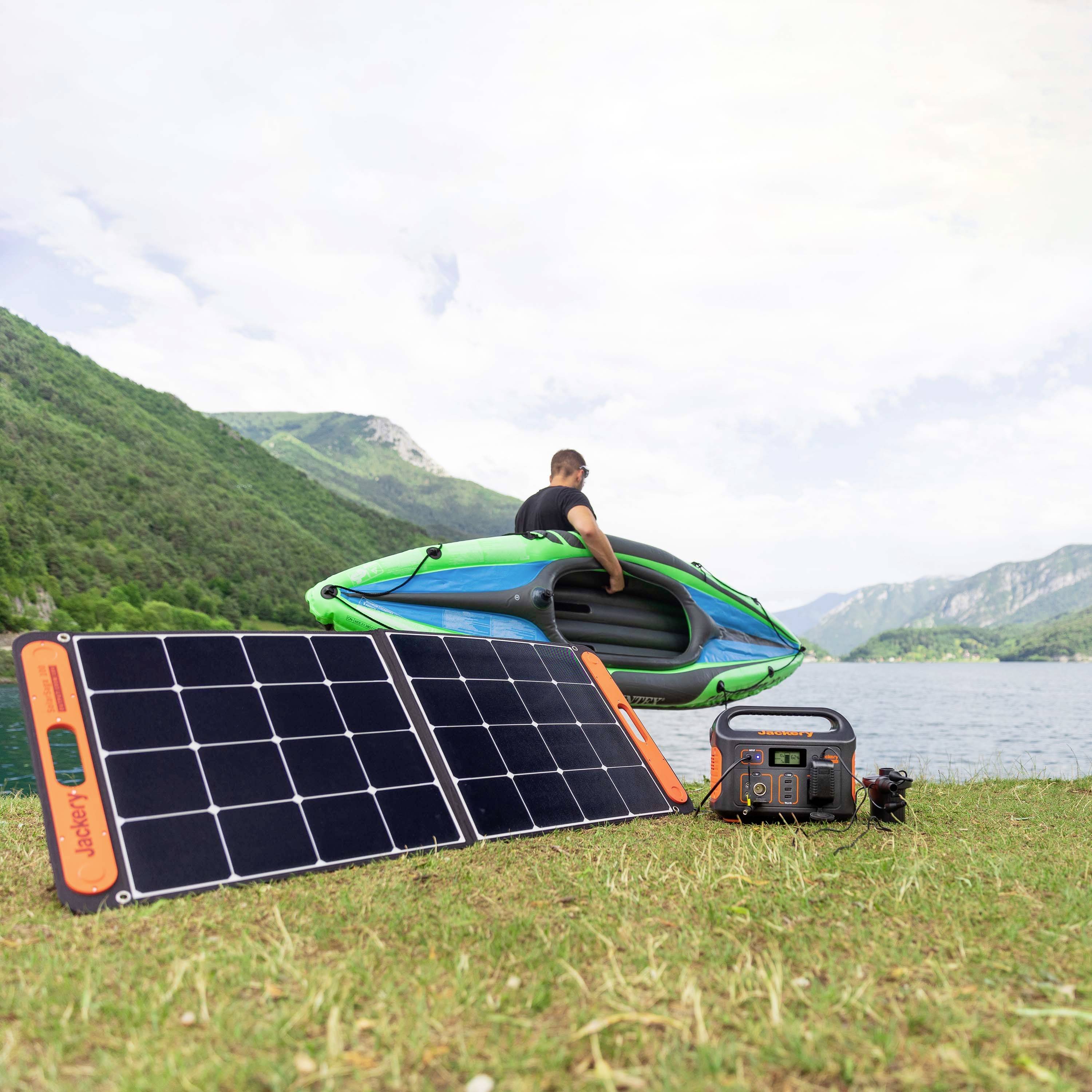 A person is lifting a kayak on the shore of a lake. In the foreground, solar panels and a portable battery device are placed on the grass.