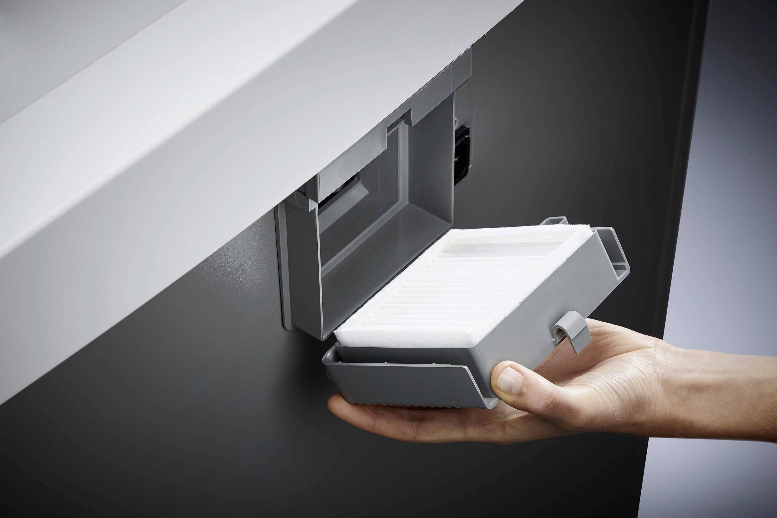 A hand removes a rectangular filter from a device, possibly an air purifier or vacuum cleaner, for cleaning.