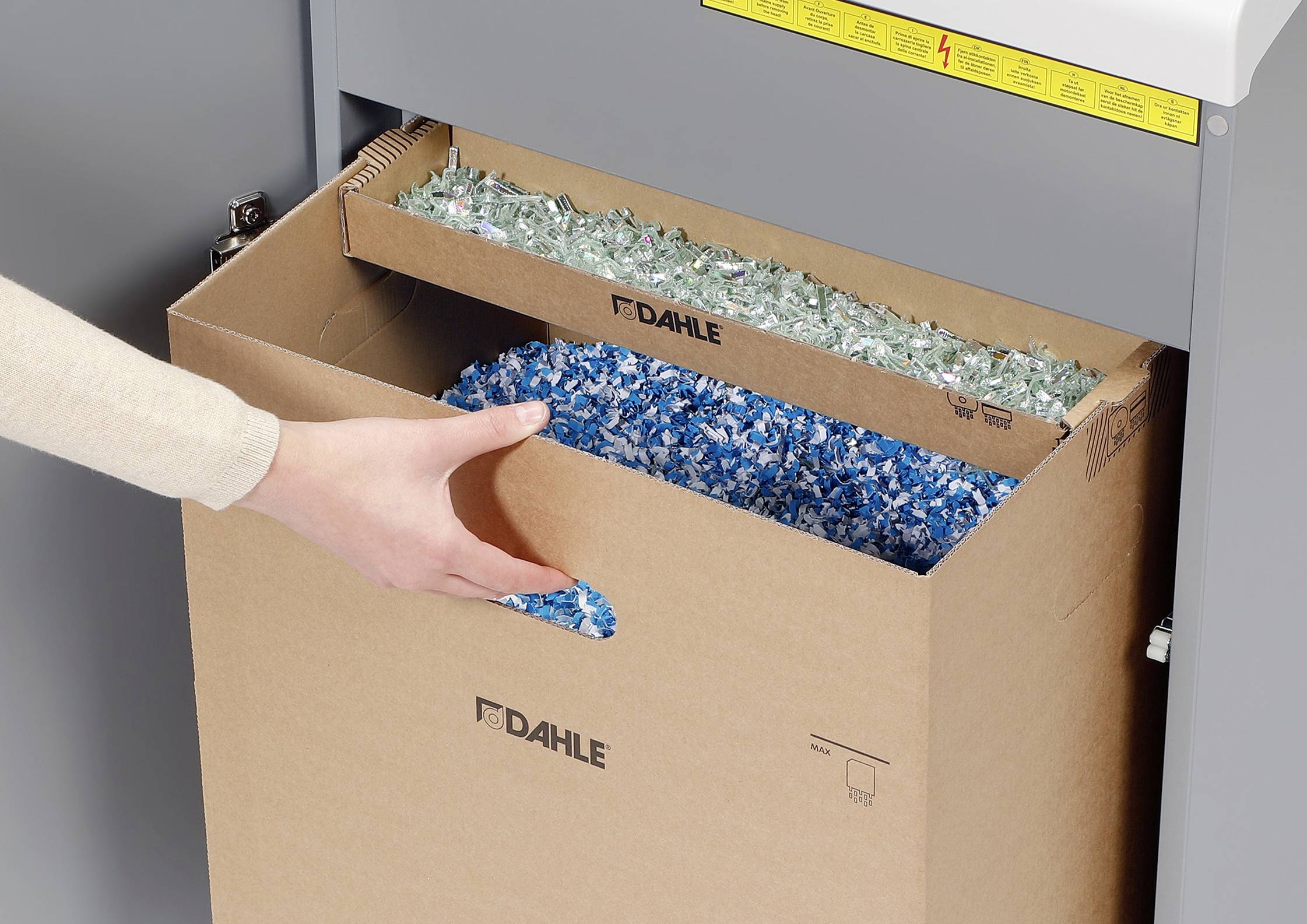 A paper shredder cuts paper into small pieces. A hand reaches for the blue and green shredded paper strips in a collection box.