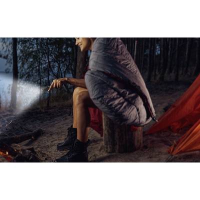 A person sits in a sleeping bag beside a campfire in a forest, holding a torch and smiling. A tent and trees are in the background.