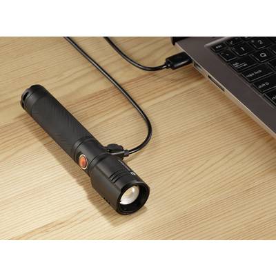 A torch is connected to a laptop with a charging cable on a wooden table.