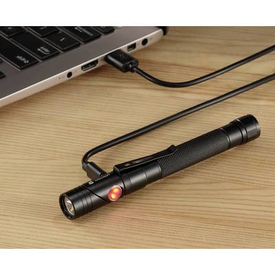A black torch is lying on a wooden-coloured table, connected to a laptop via a USB cable, with a red light illuminated on the torch.