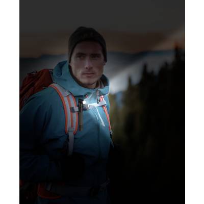 A man wearing outdoor clothing and carrying a rucksack stands in a mountainous landscape at dusk. He gazes attentively into the distance.