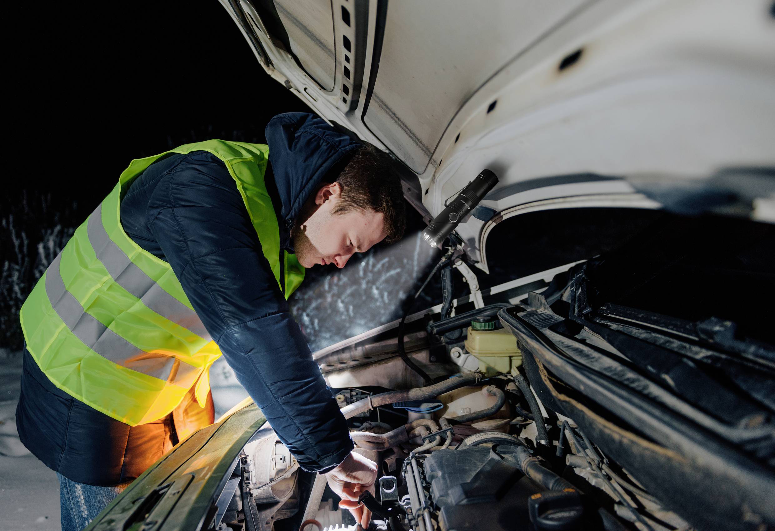 A man in a high-visibility vest is repairing a car at night, with its bonnet open. A torch illuminates the engine compartment.