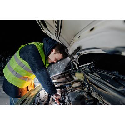 A man in a high-visibility vest is repairing a car at night, with its bonnet open. A torch illuminates the engine compartment.
