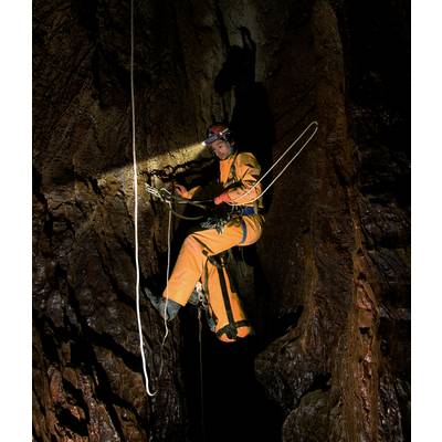 A person wearing climbing equipment descends a dark cave using ropes. A helmet with a head torch illuminates the rocky background.