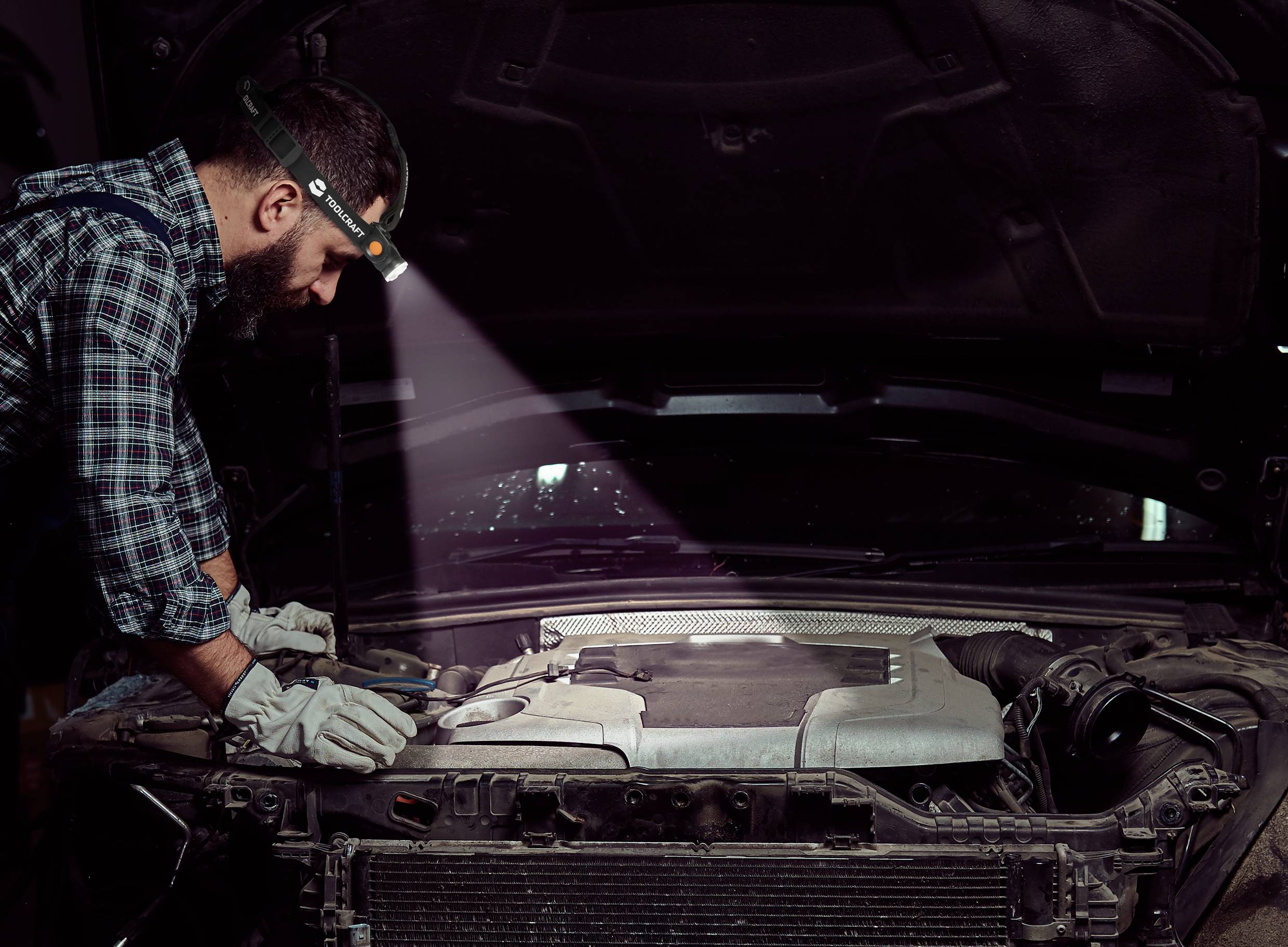 A mechanic with a head torch is working beneath an open bonnet. He is wearing gloves and inspecting the engine of a car.