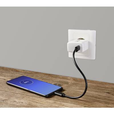 A smartphone is lying on a wooden table, charging while connected by a cable to a white socket on a grey wall.